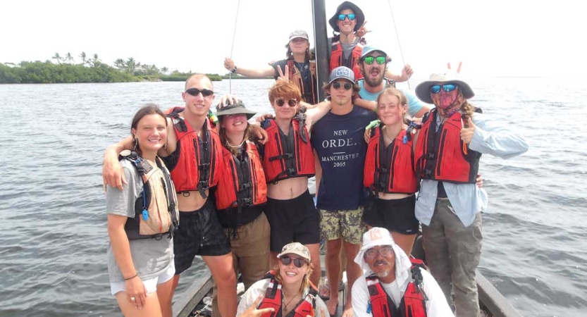 A group of students wearing lifejackets stand on a sailboat and smile for a group photo